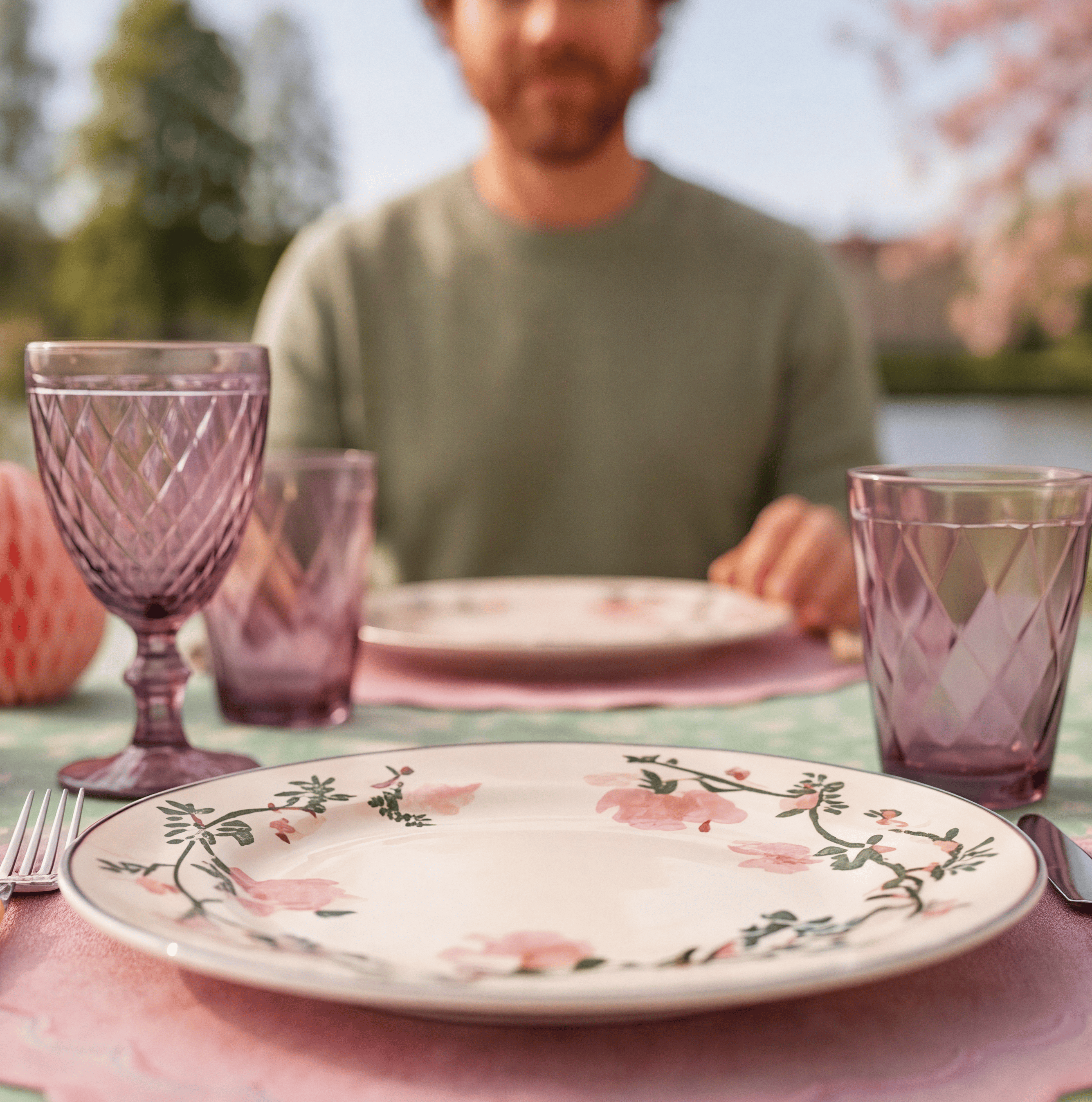 Close-up of a floral-patterned plate and purple glassware, with someone seated at the table in soft focus behind.